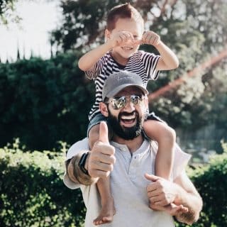 A smiling father with a beard and sunglasses carries his son on his shoulders in a sunny outdoor setting. The boy, wearing a striped shirt, playfully makes faces.