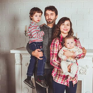 Family portrait in front of a white brick wall. A man holds a young boy while a woman holds a baby. Consider pressure washing to keep your family photos looking their best.