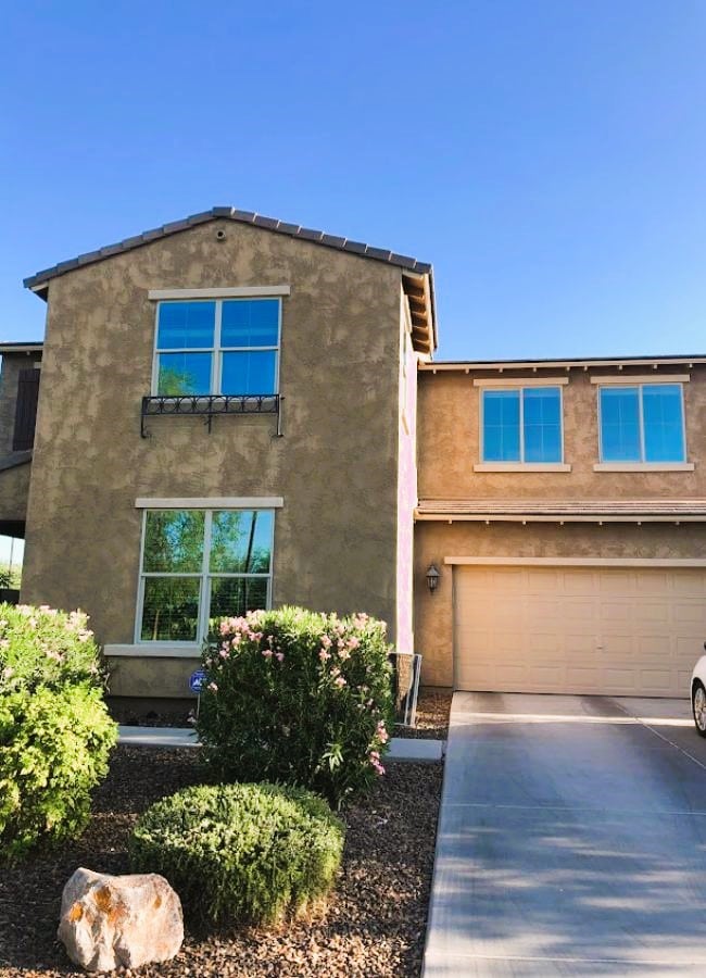 A two-story home with a stucco exterior, reflecting blue sky in its windows, sits on a well-maintained lot with desert landscaping. Regular exterior cleaning helps maintain the home's curb appeal.