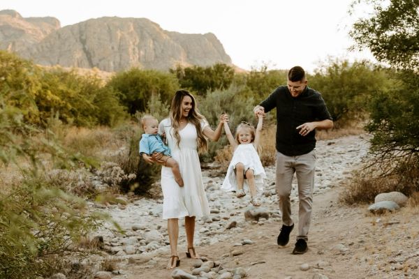 A family of four walks along a rocky path with mountains in the background. The parents hold hands with their young children, who are smiling. The mother wears a white dress, and the father wears a dark shirt and light pants.