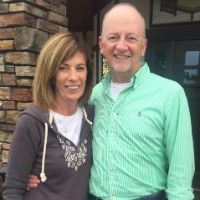 A couple smiles outdoors, possibly discussing exterior cleaning for their home. The woman wears a grey hoodie, the man a green striped shirt. Stone building in the background.