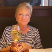 A woman with short blonde hair smiles while holding a glass of white wine. She is wearing a striped shirt and is sitting at a table in a restaurant. Consider window cleaning in Gilbert, AZ, for a crystal-clear view.