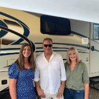 Three people stand in front of a large RV. The woman on the left wears a blue floral dress, the man in the middle wears sunglasses and a white shirt, and the woman on the right wears a green shirt and jeans. Could be related to window cleaning services.