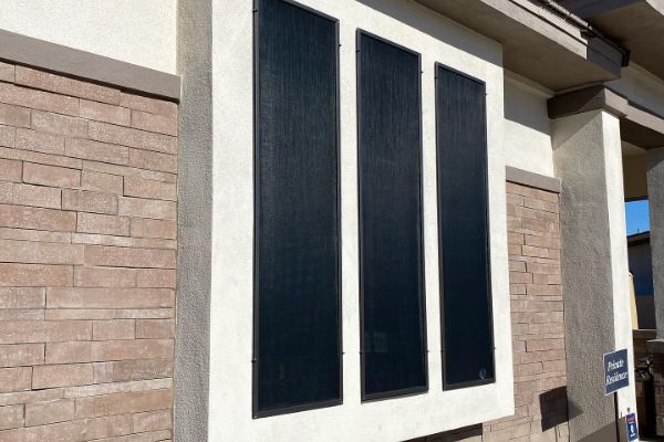 Three dark sun screens installed on a beige stucco home in Gilbert, AZ. A brick facade complements the sun screen installation, enhancing energy efficiency and curb appeal.