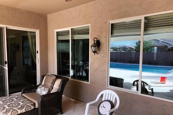 Outdoor patio with comfortable seating, white-framed sliding glass doors reflecting a pool and mountains. A white resin chair and outdoor clock add to the relaxed backyard gallery setting.