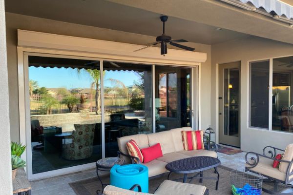 Covered patio with tan stucco walls, sliding glass doors, and a ceiling fan. Outdoor furniture includes a beige sofa with colorful pillows, a coffee table, and chairs, creating a comfortable outdoor living space.