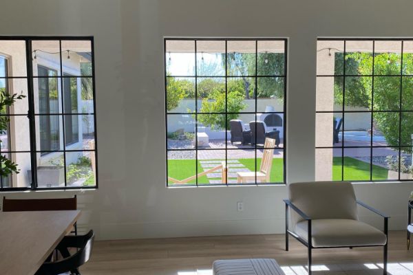 Bright, modern living space with three large windows framing an outdoor patio. Black-framed windows offer views of green lawn, patio furniture, and landscaping. Interior features light wood floors and neutral-toned furniture.