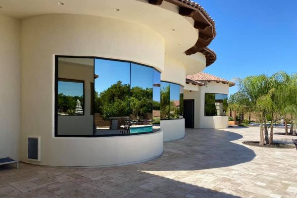 Modern curved house exterior with tinted windows reflecting the blue sky and palm trees. The home features a tiled patio and a light stucco finish.