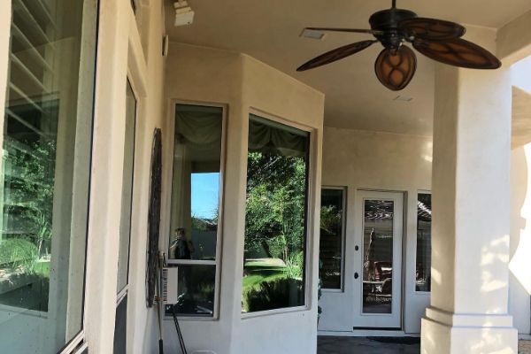 Covered patio with stucco walls, large windows reflecting green lawn, and a ceiling fan. Sunlight streams in, highlighting the architectural details.
