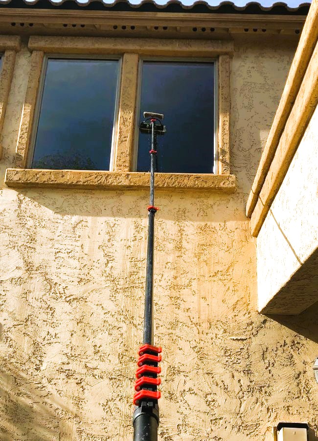 Window cleaning in Gilbert, AZ, is shown with a long pole reaching up to clean a second-story window on a stucco house with tan trim.
